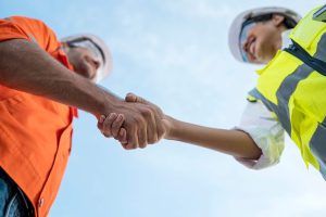 Low-angle view of two construction professionals in high-visibility safety vests and hard hats shaking hands against a clear blue sky