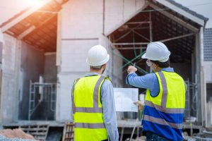Two construction professionals in high-visibility safety vests and hard hats stand with their backs to the camera, looking toward a residential building under construction while one points at a specific structural detail.