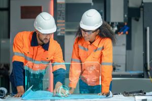 Two construction professionals in high-visibility safety gear and hard hats review a blueprint on a workbench with a glowing blue digital HUD overlay displaying technical data and 3D schematics.